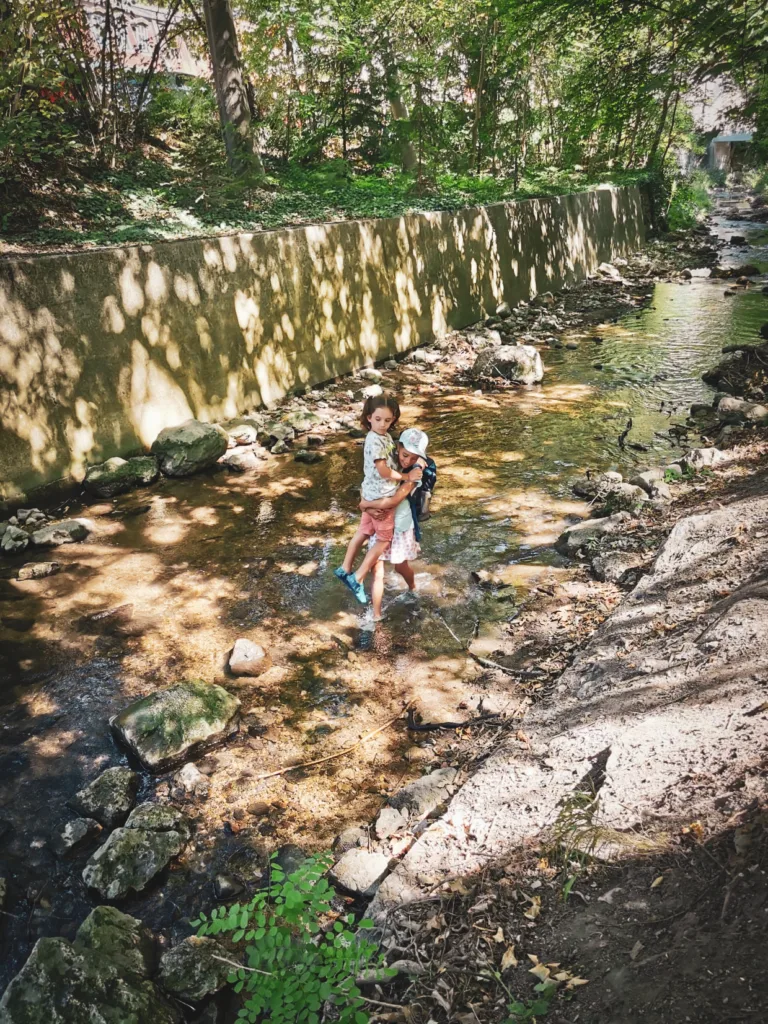 Abenteuer am Bach – kleine Entdecker genießen die Natur in vollen Zügen. Zwei Kinder waten durch einen flachen Bach, umgeben von grünen Bäumen und einem Betonufer. Sonnenlicht bricht durch die Blätter und reflektiert auf dem Wasser.
