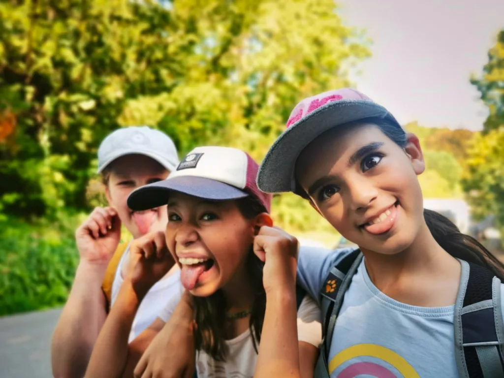 Drei Kinder mit Baseballkappen machen lustige Grimassen, strecken die Zungen heraus und halten sich die Ohren. Im Hintergrund unscharf grüne Bäume und ein sonniger Tag.