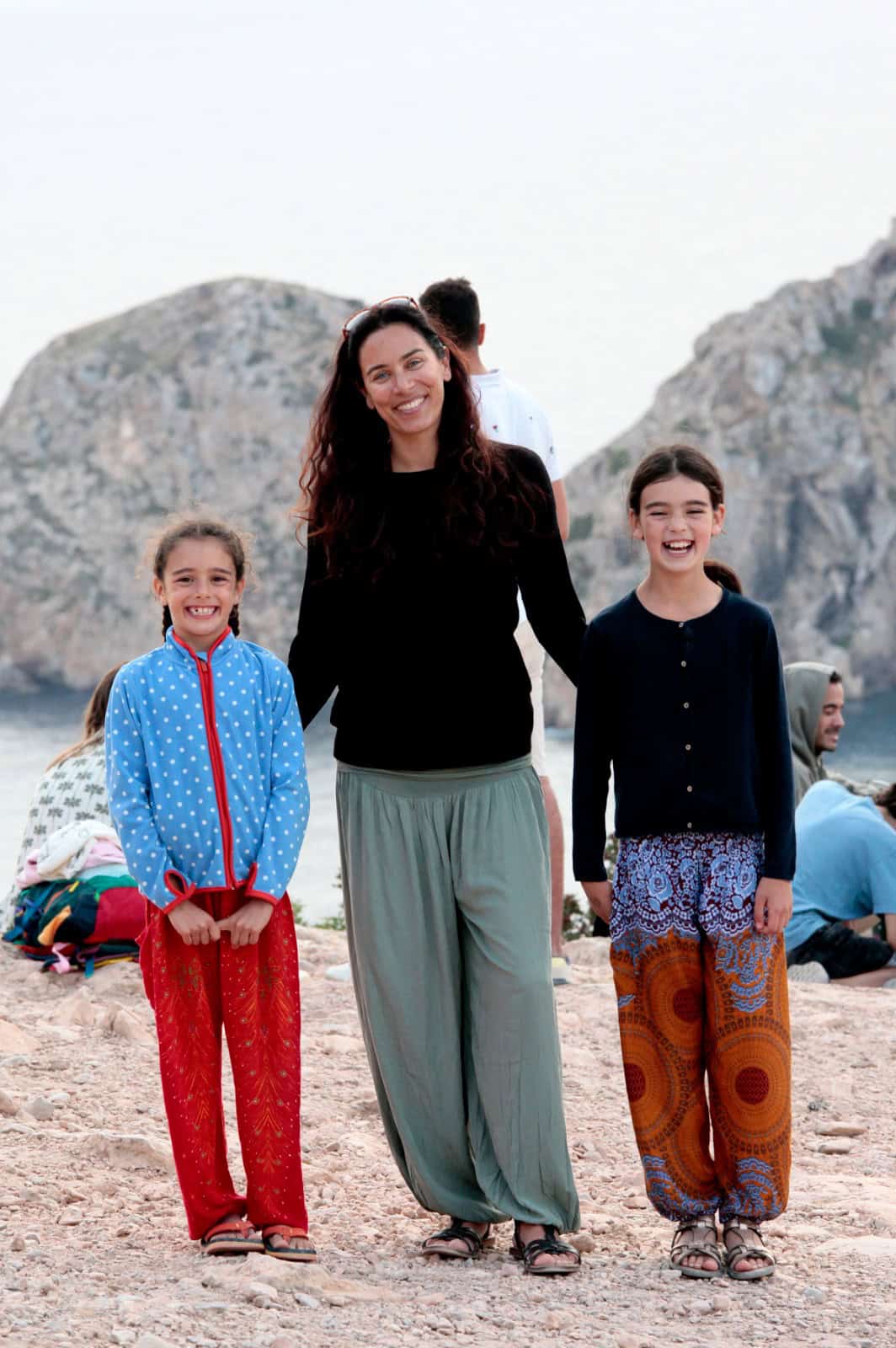 Eine Frau mit langen, dunklen Haaren steht lächelnd zwischen zwei jungen Mädchen. Alle tragen lockere, bunte Kleidung und lächeln in die Kamera. Im Hintergrund sind felsige Klippen und das Meer zu sehen.