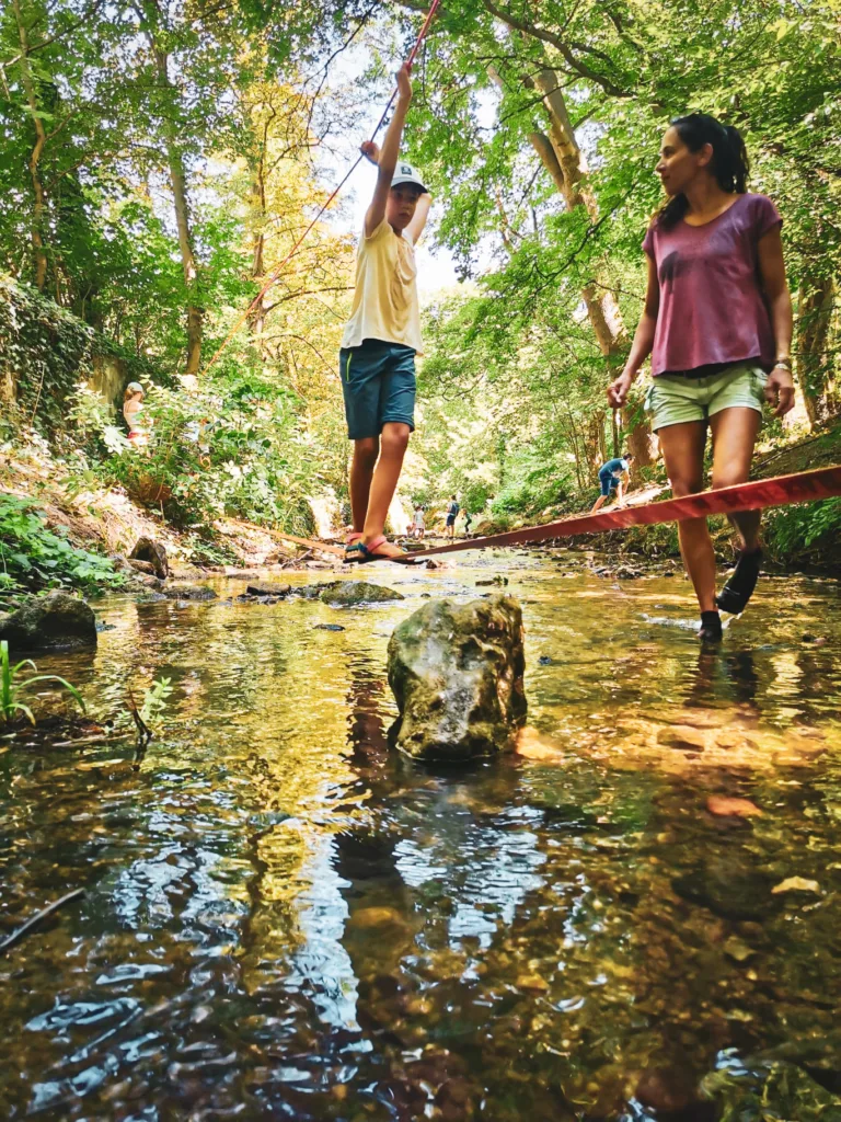 Ein Kind balanciert über eine Slackline, die über einen flachen Bach gespannt ist, begleitet von einer Frau, die aufmerksam daneben steht. Im Hintergrund dichte grüne Bäume und weitere Kinder beim Spielen.