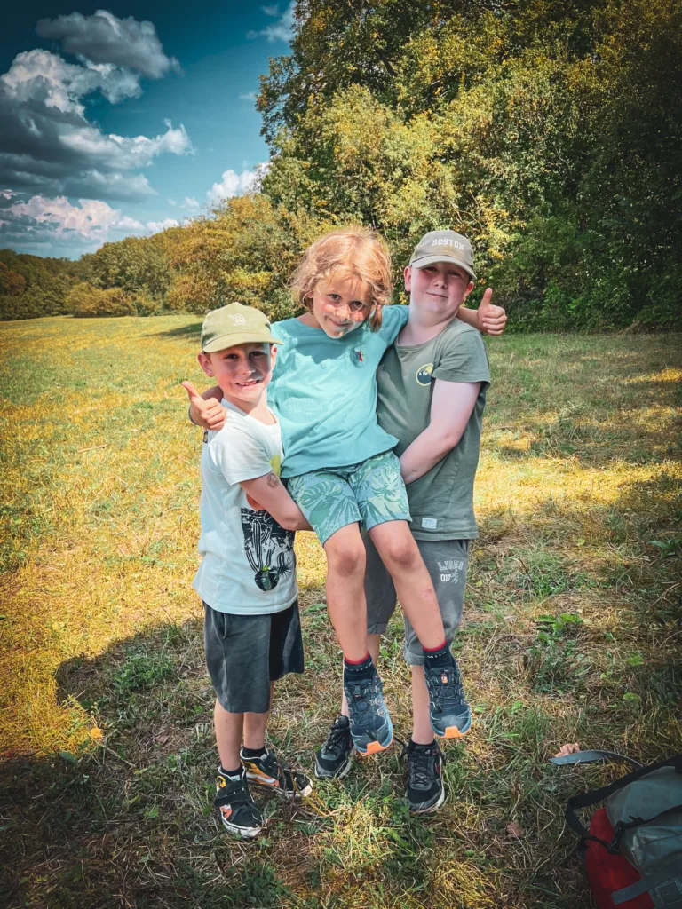 Drei Kinder stehen auf einer Wiese im Abenteuercamp, zwei von ihnen heben lachend das dritte Kind hoch, während sie von Bäumen umgeben sind und die Sonne scheint.