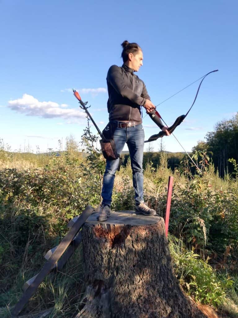 Ein Mann mit schwarzer Jacke steht auf einem großen Baumstumpf und spannt einen traditionellen Bogen. Die Szene spielt sich in einer natürlichen Umgebung mit Wiesen und Bäumen im Hintergrund bei klarem Himmel ab.