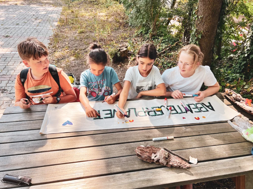 Vier Kinder sitzen an einem Holztisch im Freien und gestalten gemeinsam ein Banner mit der Aufschrift 'ABENTEUERPLANET'. Sie verwenden bunte Marker und dekorieren es mit Herz- und Blumenmotiven.