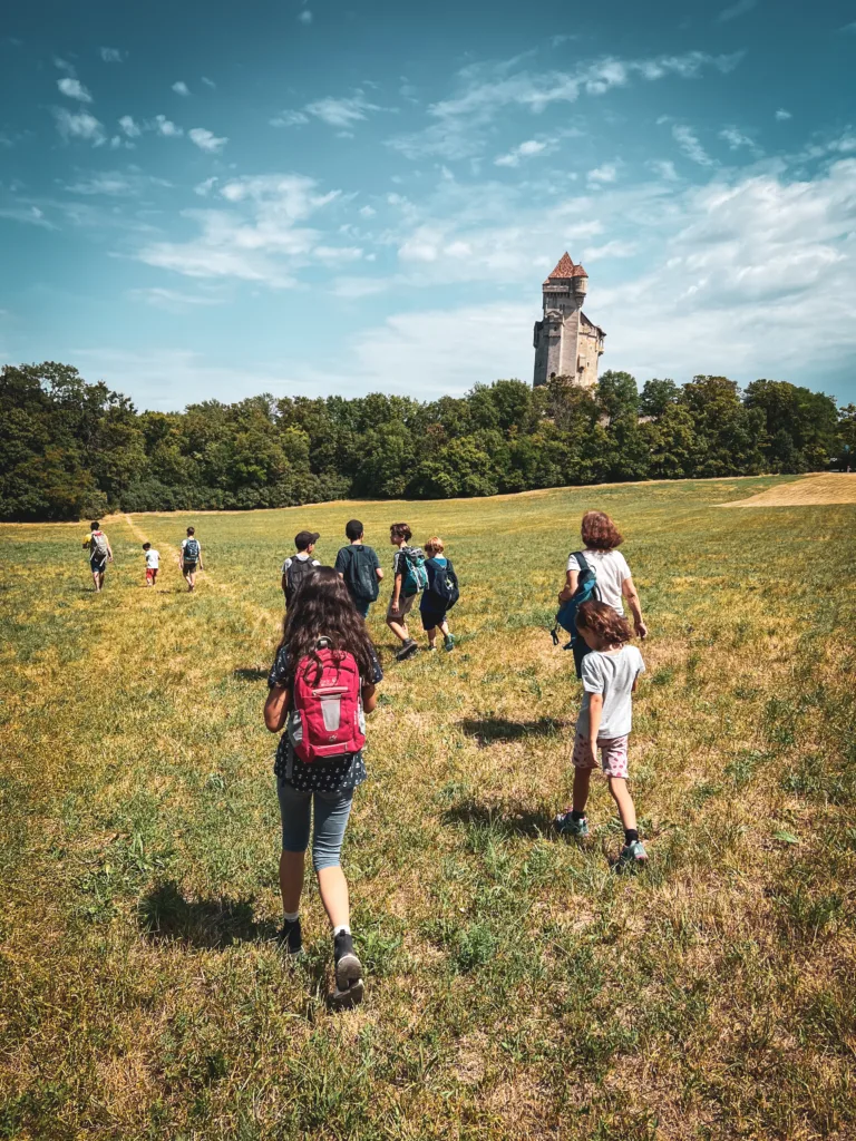 Eine Gruppe von Kindern wandert über eine Wiese in Richtung einer mittelalterlichen Burg, die von Bäumen umgeben ist, unter einem klaren blauen Himmel.