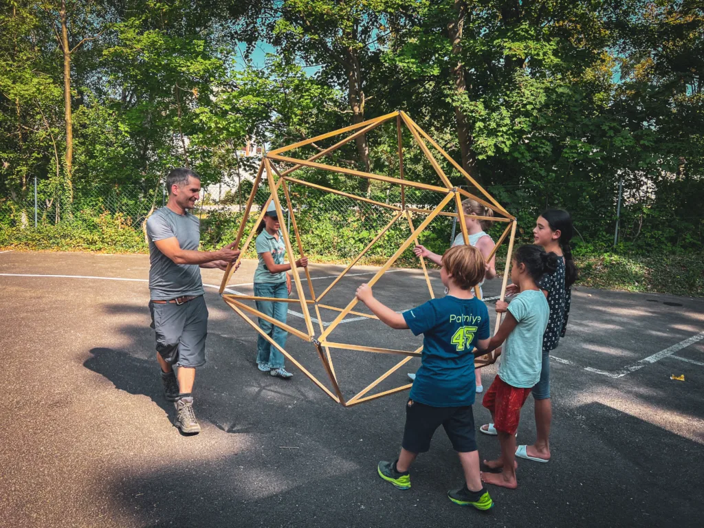 Ein Betreuer und eine Gruppe von Kindern halten und bewegen gemeinsam eine große geometrische Holzkonstruktion auf einem Spielplatz, umgeben von Bäumen.