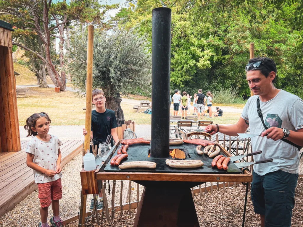 Ein Betreuer grillt Würstchen auf einer Feuerplatte, während zwei Kinder danebenstehen und zuschauen. Im Hintergrund spielen weitere Teilnehmer des Abenteuercamps auf einer Wiese.