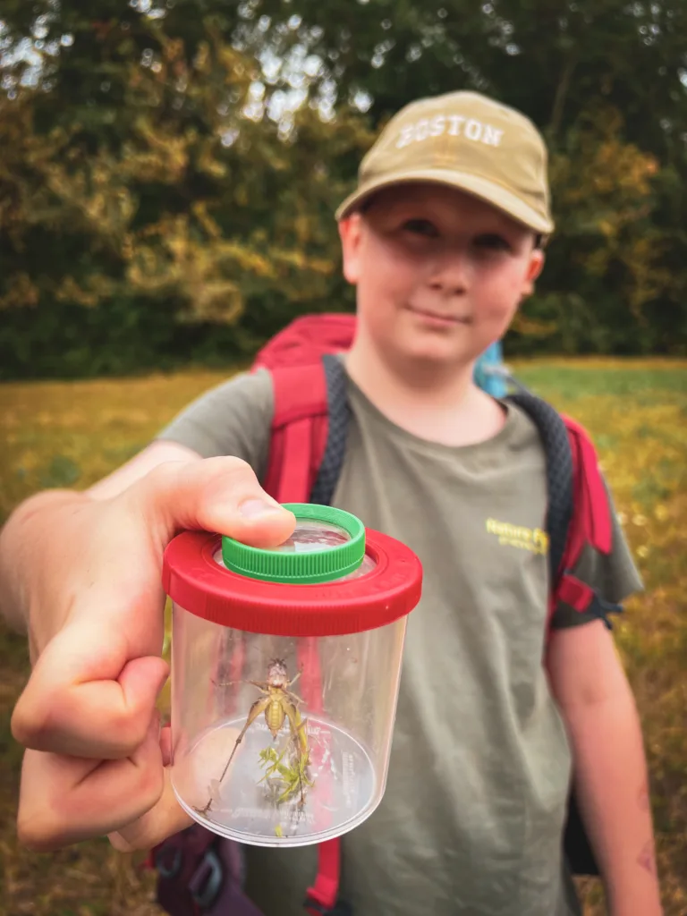 Ein Junge mit einem Rucksack und einer Baseballkappe hält stolz einen kleinen Behälter mit einer Heuschrecke, die er in der Natur gefangen hat.