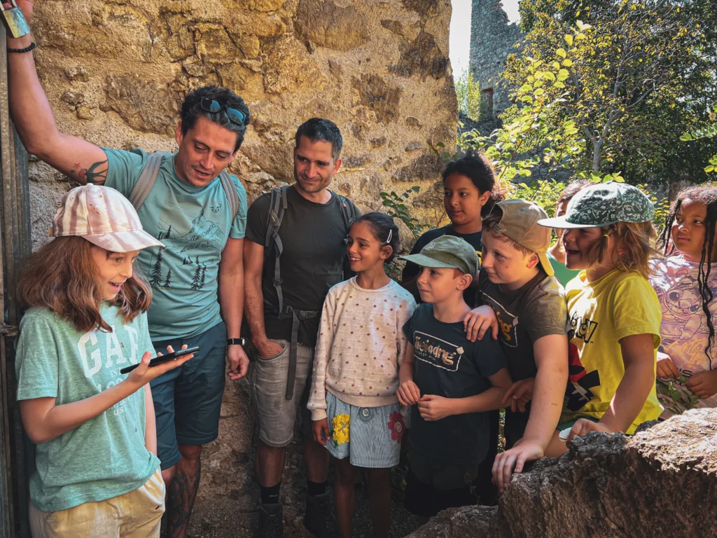 Eine Gruppe von Kindern und Betreuern steht in einer historischen Ruine, während ein Mädchen etwas auf einem Smartphone präsentiert. Alle hören aufmerksam zu und lächeln.