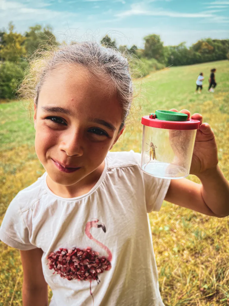Ein Mädchen in einem weißen T-Shirt mit Flamingo-Design hält einen Behälter mit einer Heuschrecke und lächelt in die Kamera, im Hintergrund eine grüne Wiese mit spielenden Kindern.
