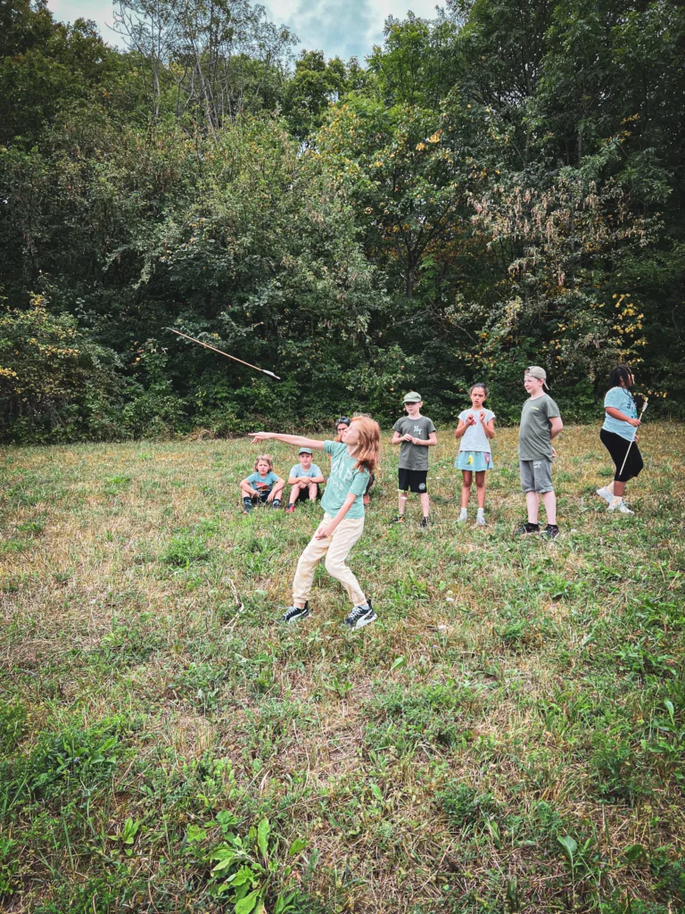 Ein Mädchen wirft einen Pfeil auf einer Wiese, während andere Kinder zuschauen oder sitzen, mit einem dichten Wald im Hintergrund.