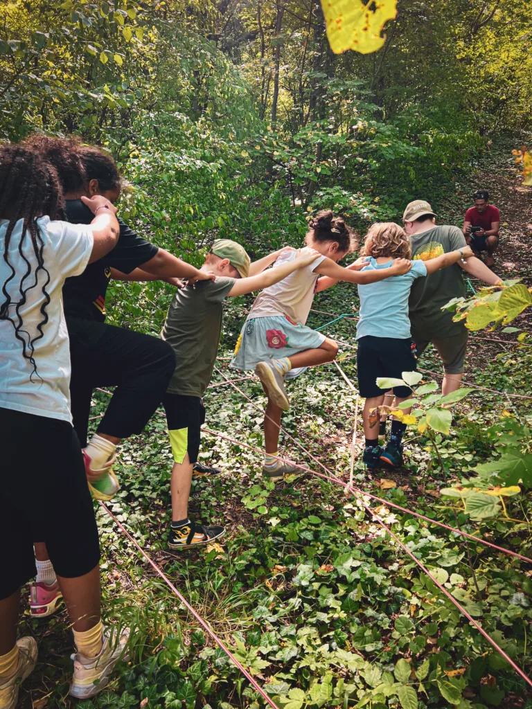 Eine Gruppe von Kindern balanciert gemeinsam über gespannte Seile in einem Wald, wobei sie sich gegenseitig an den Schultern festhalten.