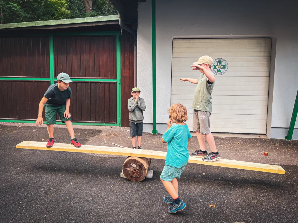 Vier Kinder spielen auf einer selbstgebauten Wippe aus einem Holzbrett und einem Baumstamm, während sie Gleichgewicht und Geschicklichkeit üben.