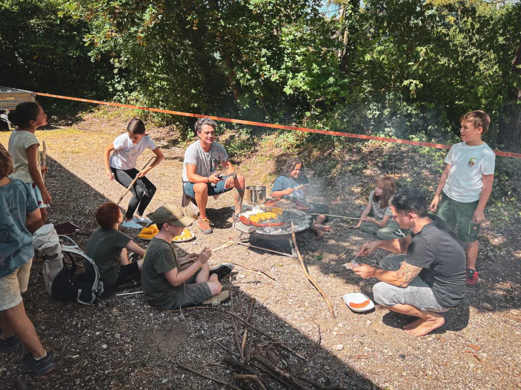 Eine Gruppe von Kindern und Betreuern sitzt um ein Lagerfeuer, grillt Würstchen und Brot auf Stöcken und genießt das gemeinsame Outdoor-Kochen.