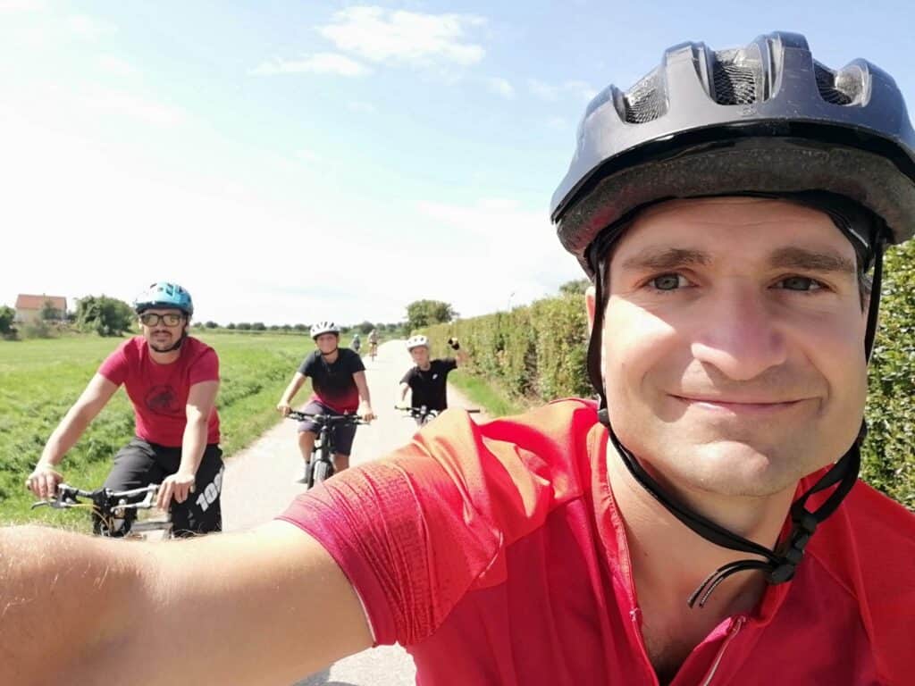 Eine Gruppe von vier Fahrradfahrern, alle mit Helmen, fährt auf einem sonnigen Weg durch eine ländliche Gegend mit grünen Wiesen und Hecken. Im Vordergrund macht ein Mann im roten Trikot ein Selfie, während er auf seinem Fahrrad fährt.