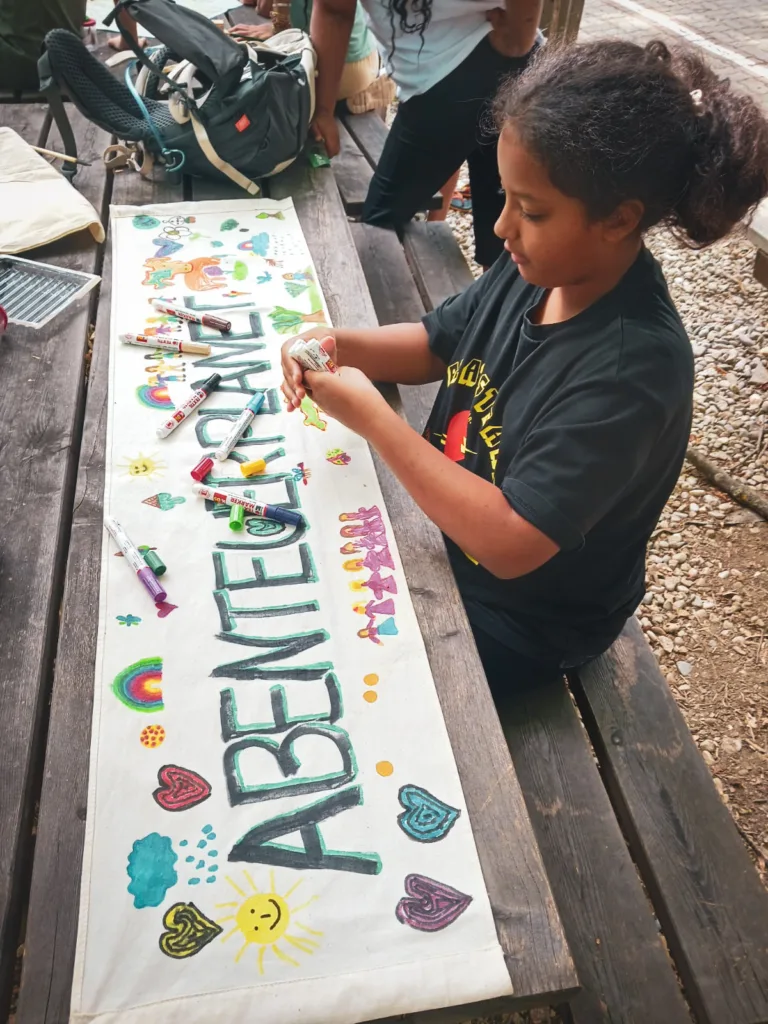 Ein Kind sitzt an einem Holztisch und dekoriert ein Banner mit dem Wort 'ABENTEUERPLANET', umgeben von bunten Zeichnungen wie Herzen, Sonnen und Regenbögen. Filzstifte liegen verstreut auf dem Tisch