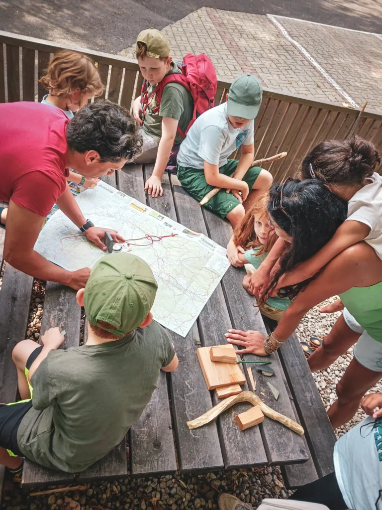 Eine Gruppe von Kindern und ein Betreuer sitzen um einen Holztisch und betrachten gemeinsam eine Karte, umgeben von Werkzeugen und geschnitzten Holzstücken.