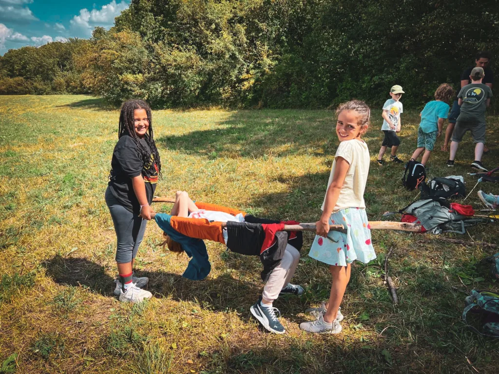 Zwei Mädchen tragen lachend ein improvisiertes Tragegestell aus Ästen und einer Jacke auf einer Wiese, während weitere Kinder im Hintergrund spielen.