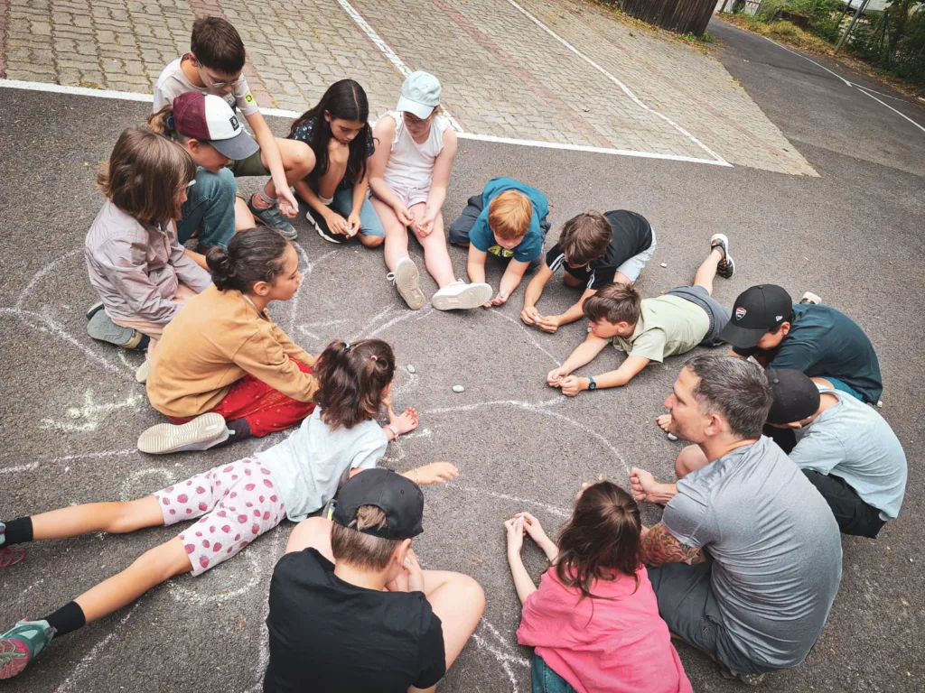Eine Gruppe von Kindern und einem Erwachsenen sitzt im Kreis auf einem asphaltierten Platz und spielt ein Kreisspiel mit kleinen Steinen.